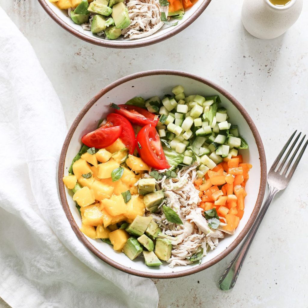 Two bowls of shredded chicken mango salad with tomatoes, orange bell peppers, avocado and diced cucumbers.