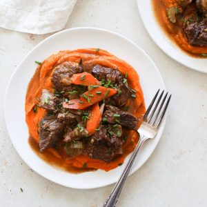 Braised beef on a plate of orange mashed sweet potatoes with carrots and chives.