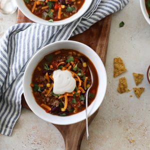 Two bowls of taco soup with shredded cheese, a dollop of cream, cilantro, and a side of crushed chips.