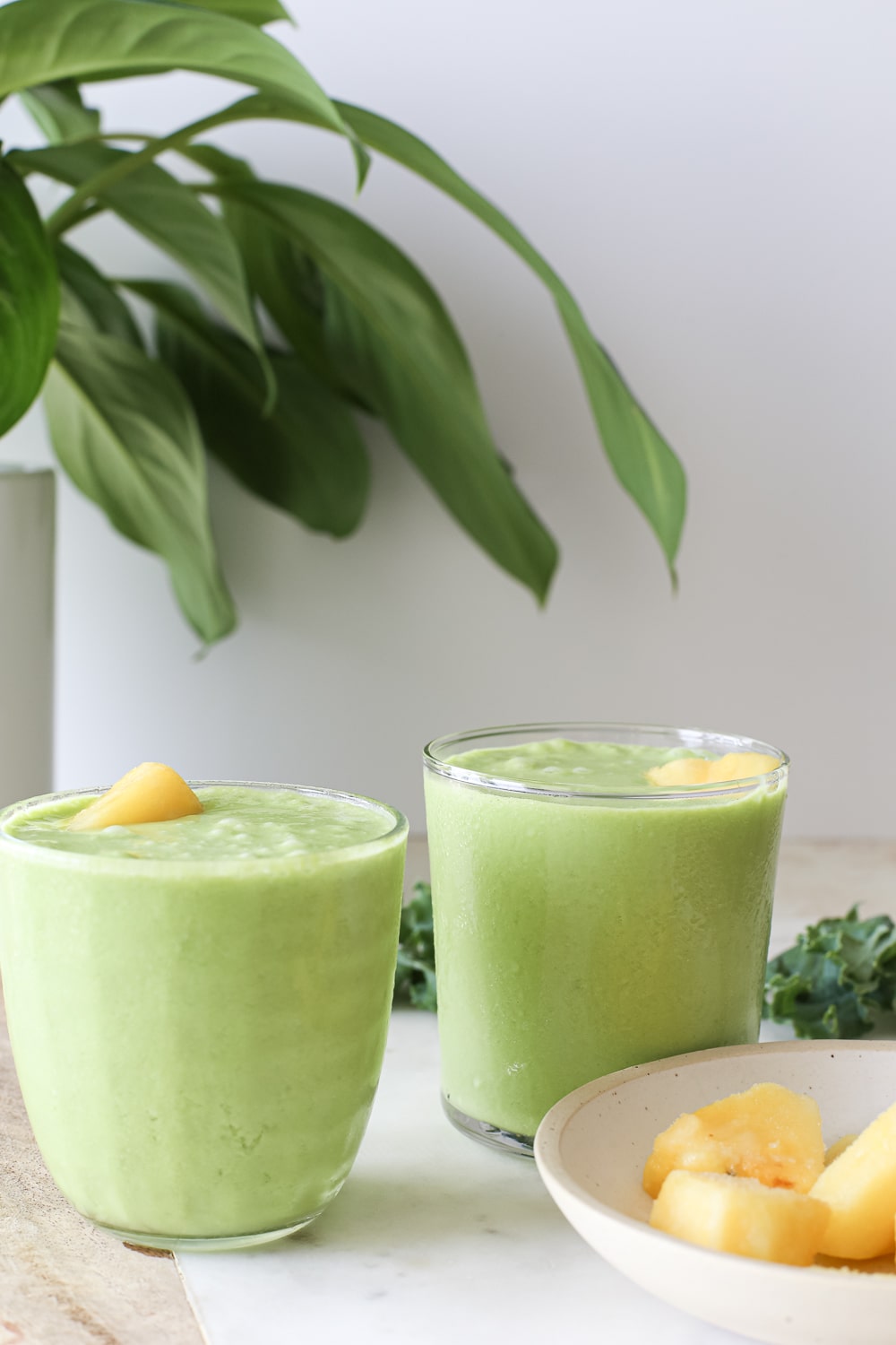 two glasses of green piña colada smoothies next to a bowl of frozen pineapple chunks and a green plant in the background