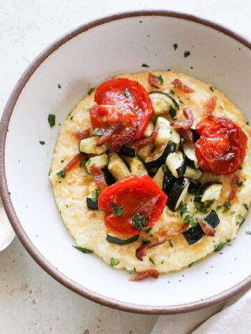 A bowl of savory breakfast polenta with roasted tomatoes, zucchini, crispy bacon bits, and fresh herbs.