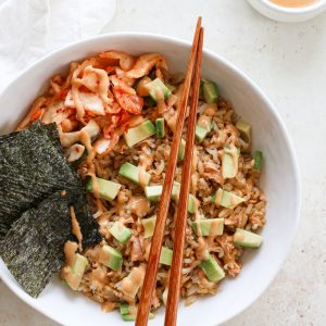 Large salmon rice bowl with chopsticks topped with avocado, kimchi, and nori with a side of spicy mayo.