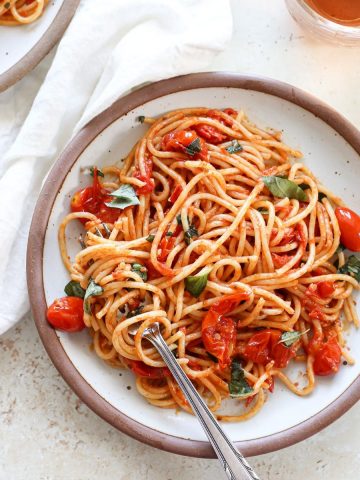 A plate of spaghetti noodles with small burst cherry tomatoes in a sauce with fresh basil.