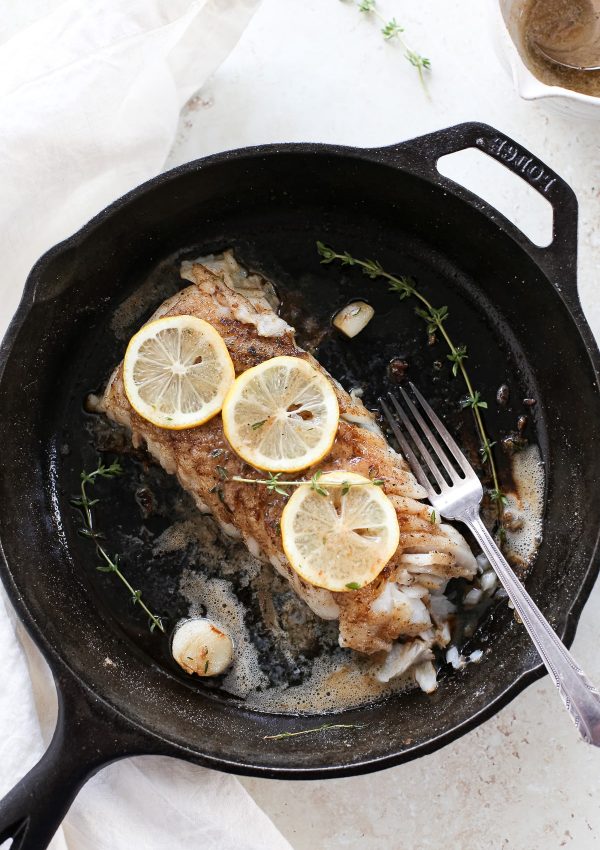 A cooked fillet of lemon butter fish in a skillet with fresh lemon slices and a fork.