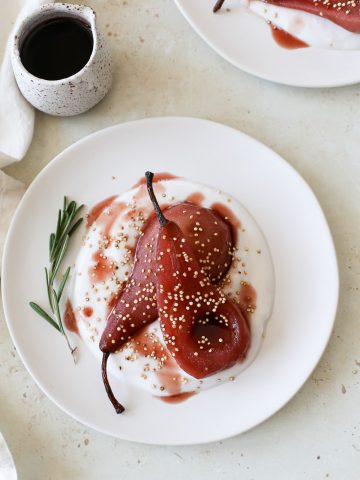 A beautiful plate of deep red poached pear halves on a bed of yogurt with syrup drizzled, a sprinkle of toasted quinoa, and a sprig of rosemary.