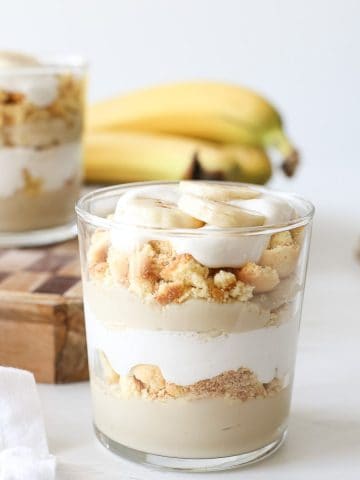 Two glasses with layers of dairy-free banana pudding, crushed vanilla wafers, dairy-free coconut whipped cream, and fresh banana slices.
