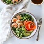 Shrimp poke bowls with sushi rice and colorful fresh vegetables such as ribboned carrots, sliced avocado, arugula, sugar snap peas, and cucumber.