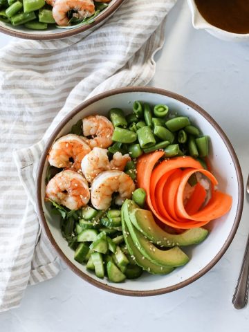 Shrimp poke bowls with sushi rice and colorful fresh vegetables such as ribboned carrots, sliced avocado, arugula, sugar snap peas, and cucumber.