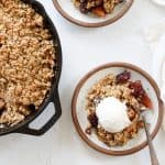 Two plates with apple and blackberry crumble with a scoop of vanilla ice cream on top next to a skillet of crumble.