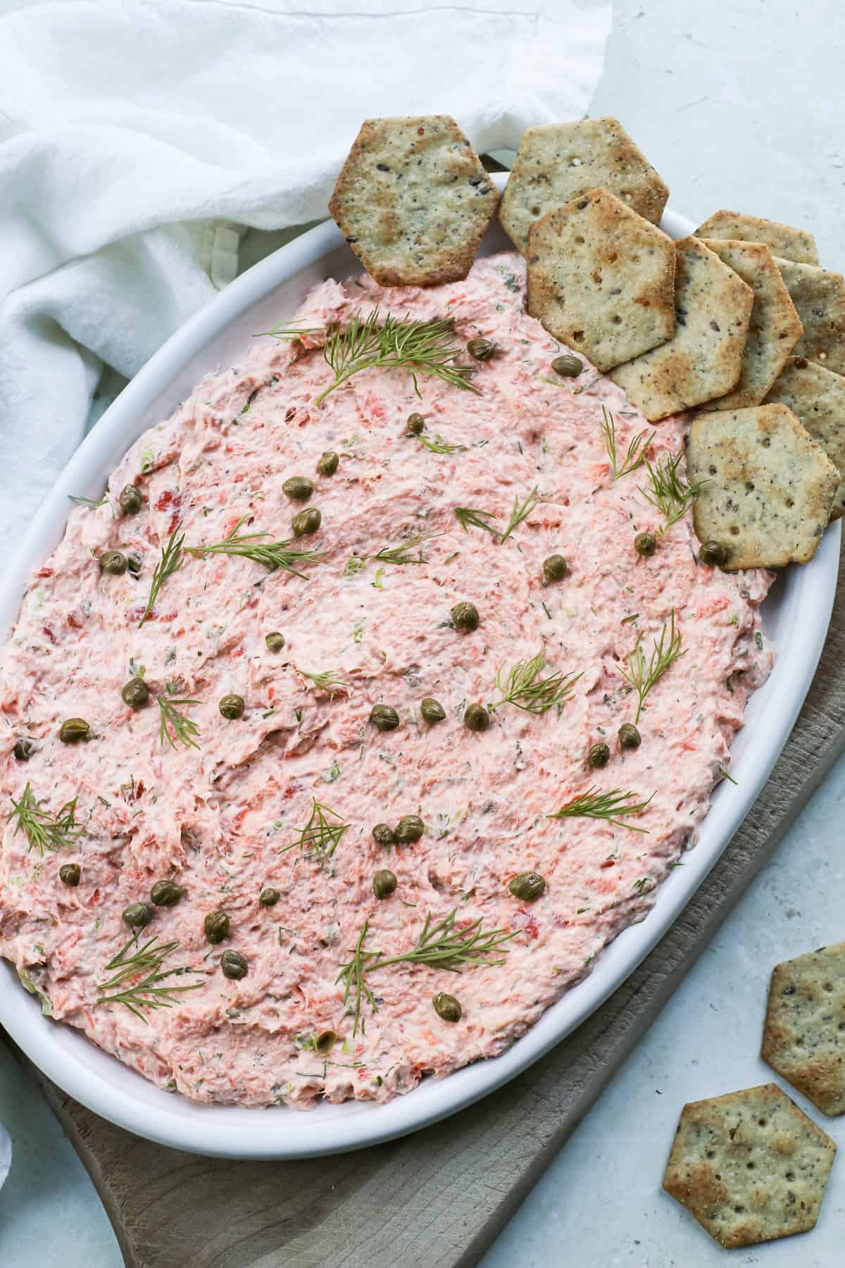 An oval platter with smoked salmon dip without cream cheese topped with fresh dill sprigs, capers, and crackers.