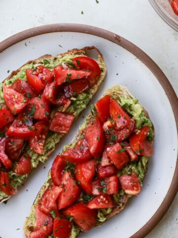 Two pieces of avocado toast on sourdough bread with marinated tomatoes and fresh parsley.
