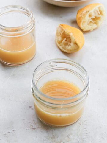 Two small glass jars of lemon ginger shots next to a bowl of juiced lemon halves.