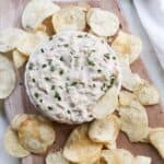 A bowl of dairy free french onion dip with chives on a cutting board with potato chips.
