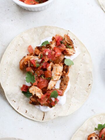 Chopped pieces of adobo chicken layered on on a corn tortilla with a smear of plain yogurt, topped with pico de gallo, and cilantro leaves next to a bowl of lime slices, and more of the toppings in bowls.