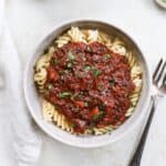 A bowl of fusilli pasta with a generous mound of a hearty lentil bolognese red sauce with minced parsley next to a fork.