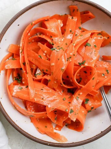 A white bowl with a brown rim with orange carrot ribbons tossed in a dressing and topped with minced parsley and hemp seeds.