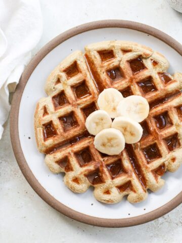 A waffle on a white plate with a brown rim topped with fresh banana slices next to a small ceramic jar of real maple syrup.