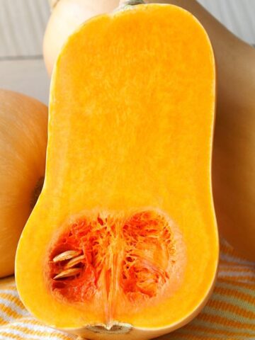 A butternut squash sliced in half with the seeds and orange flesh exposed leaning against other whole butternut squash.