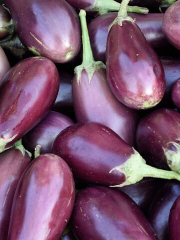 A pile of medium purple eggplants (also called aubergines) with green stems.